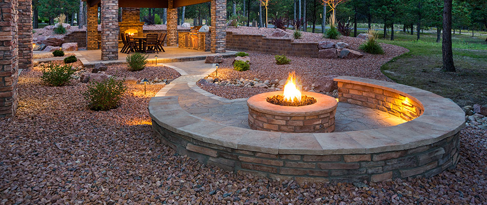 A fire pit with matching seating wall lit up in the evening surrounded by extravagant landscaping in Ankeny, IA.